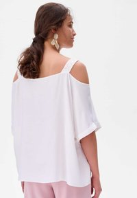 Biała bluzka cold-shoulder o luźnym kroju, z krótkimi rękawami i dekoracyjnymi paskami. Gładka faktura z delikatnym połyskiem, zestawiona z jasnoróżowymi spodniami.