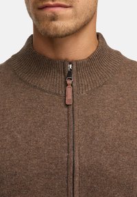 Pull en laine marron avec un col haut, doté d'une fermeture éclair métallique et d'une tirette en cuir. La texture est douce et finement côtelée.