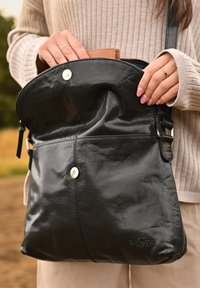Bolso de cuero negro con un acabado texturizado, cierre con cremallera, botones de presión y una correa ajustable. Una cartera parcialmente visible en el interior.