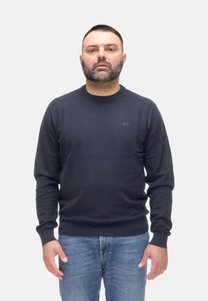 Uomo con barba e capelli corti, indossa un maglione blu scuro e jeans blu, in piedi davanti a uno sfondo grigio chiaro uniforme.