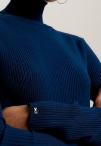 Persoon draagt een blauwe geribbelde coltrui met lange mouwen en zilveren oorringen, armen gekruist tegen een neutrale achtergrond.