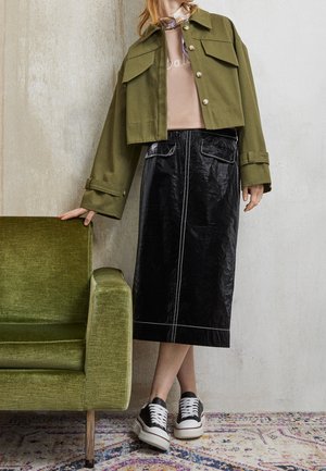 Personne portant une veste olive courte, un haut beige, une jupe midi noire et des baskets noir-blanc se tenant à côté d'un fauteuil vert sur un tapis à motifs.