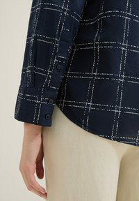 Chemise bleu marine avec motif en damier blanc, poignets à boutons, tissu doux, ourlet légèrement arrondi, portée avec un pantalon clair.