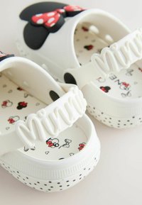 Zapatos blancos sin cordones con orejas de ratón negras y diseño de lazo rojo, decorados con el texto "Minnie" en la tira y patrones de Minnie Mouse en el interior.