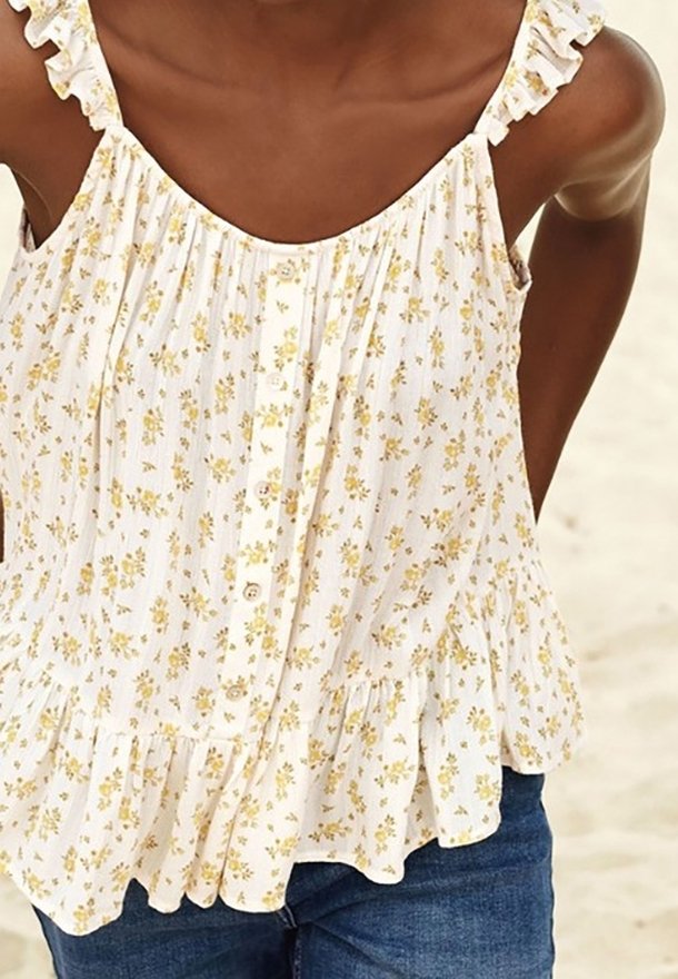 Blouse blanche sans manches avec de petites fleurs jaunes, boutons à l'avant, bretelles et ourlet à volants, portée avec un jean bleu.