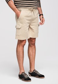 Hombre con pantalones cortos cargo beige con bolsillos laterales y zapatillas negras, de pie con una mano en el bolsillo frente a un fondo liso.