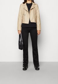 Beige faux leren bikerjack met zilveren ritsen, gecombineerd met een zwart topje en zwarte uitlopende broek met kliksluitingen en zwarte laarzen.