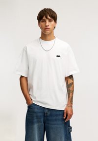 Hvid bomulds t-shirt med korte ærmer, rund hals og et lille sort logo på brystet. Båret med blå løse denim shorts.