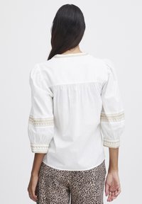 Blouse blanche avec des manches bouffantes, ornée d'accents brodés le long du col et des manches. Portée avec un pantalon à motifs.