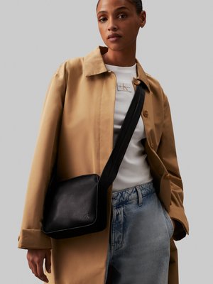 Borsa a tracolla nera in pelle con una texture liscia, piping bianco e un logo sottile. Indossata sopra un cappotto beige e jeans chiari.