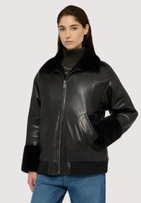 Czarna skórzana kurtka z futrzanym kołnierzem, na zamek błyskawiczny i ściągaczami przy mankietach. Ma boczne kieszenie i luźny krój, zestawiona z niebieskimi dżinsami.