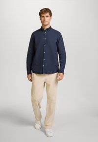 Camicia blu navy con bottoni in cotone, con colletto classico, abbinata a pantaloni beige in velluto a coste e sneaker bianche. Design semplice, senza motivi.