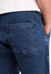 Jeans de mezclilla de color azul oscuro, con un bolsillo trasero que presenta detalles de costura y un acabado de tela texturizada.