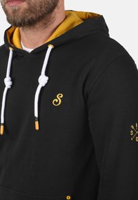 Zwarte hoodie met een gele binnenkant, witte trekkoorden en een klein geel geborduurd logo. Heeft oranje uiteinden aan de trekkoorden en details op de mouwen.
