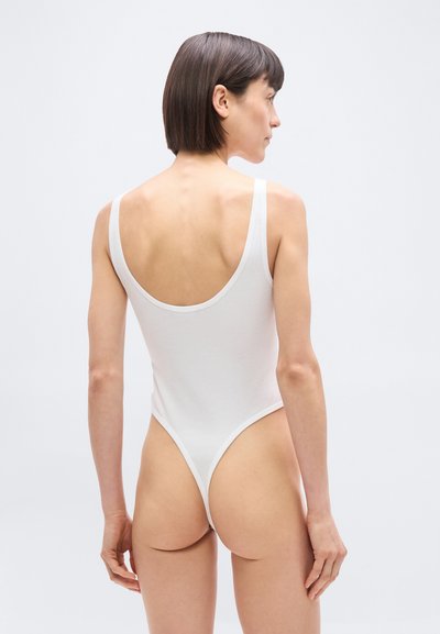 Γυναίκα με κοντά σκούρα μαλλιά, φωτογραφημένη από πίσω, φοράει λευκό κορμάκι χωρίς μανίκια σε απλό ανοιχτό φόντο.