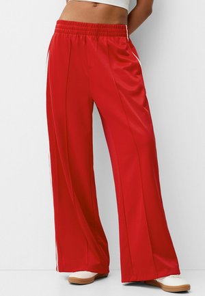 Osoba ubrana w luźne czerwone spodnie dresowe z białymi bocznymi paskami i białe trampki, stojąca na tle jednolitego białego tła.