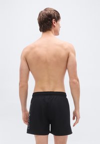 Μαύρα μαγιό σορτς με ελαστική μέση, με λεπτομέρεια λογότυπου στο πλάι. Λεία υφή, μήκος μέχρι το μέσο του μηρού, χωρίς επιπλέον σχέδια.