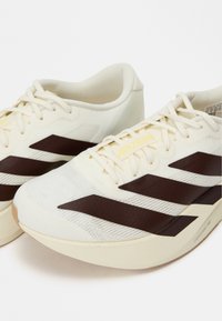 Laufschuhe mit einem weißen Mesh-Obermaterial, dunkelbraunen diagonalen Streifen und gelbem "ADIZERO"-Branding. Leichtes Design mit einer strukturierten Sohle.