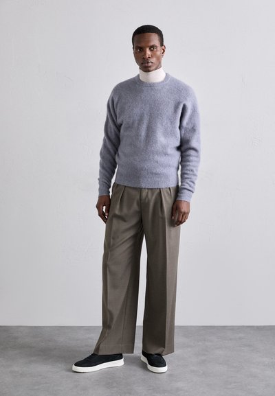 Grå, lodden genser, kremfarget turtleneck, skreddersydde brune bukser og svarte joggesko. Mannen står med avslappet holdning mot en nøytral bakgrunn.