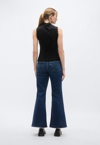 Μαύρο αμάνικο τοπ με κουμπιά στα μανίκια, συνδυασμένο με μπλε τζιν καμπάνα με πίσω τσέπες και κόκκινη ετικέτα λογότυπου.