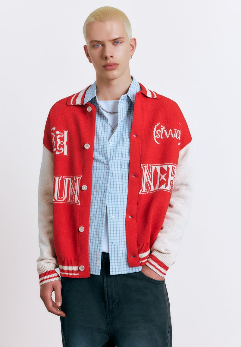 Jeune personne aux cheveux blonds courts portant une veste universitaire rouge et blanche sur une chemise à carreaux bleue et un pantalon foncé, debout avec une main dans la poche.