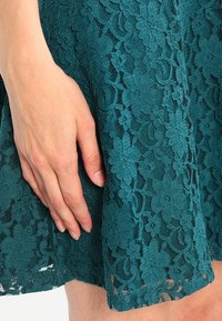 Türkiser Spitzenstoff mit floralen Mustern und einer strukturierten Oberfläche, auf dem sanft eine Hand ruht, die die Details hervorhebt.