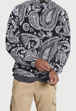 Personne portant une veste en polaire noire et blanche à motifs paisley et un pantalon cargo beige, ajustant le poignet de la veste.