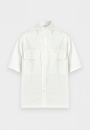 Homme aux cheveux bouclés foncés portant une chemise blanche à manches courtes boutonnée sur un tee-shirt blanc, un jean bleu, une ceinture noire et des chaussures noires, debout avec une main derrière le dos.