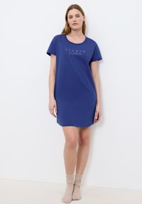 Kobieta w niebieskiej koszuli nocnej z krótkim rękawem, z nadrukiem faz księżyca, i beżowych skarpetkach, stojąca na tle białej ściany.