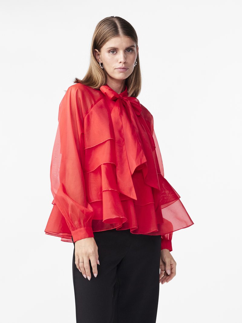 Red sheer blouse with ruffled layers, long sleeves, and a tied collar. Paired with black pants, it features a light and airy texture.