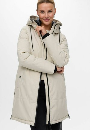Mujer con un abrigo de invierno beige claro con capucha, cremallera negra y cordones, pantalones negros, sonriendo y tocando el cuello del abrigo.