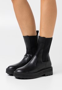Botas de tobillo de cuero negro con paneles laterales elásticos, punta redondeada y suela de goma gruesa con un patrón de banda de rodadura para mayor agarre.