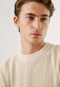 Cremfarbener, gestrickter Pullover mit ribbeligem Rundhalsausschnitt und strukturiertem Stoff. Das Design zeigt ein lockeres Strickmuster im gesamten Pullover.