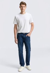Camiseta blanca de algodón para hombre combinada con vaqueros de mezclilla azul oscuro. Presenta un ajuste clásico, pierna recta y detalles sutiles de costura. Zapatillas deportivas blancas.
