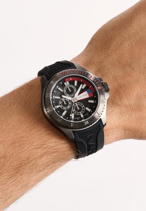 Armbanduhr mit schwarzem Zifferblatt, roten und blauen Details, drei Hilfszifferblättern und schwarzem strukturiertem Gummiband am Handgelenk einer Person.