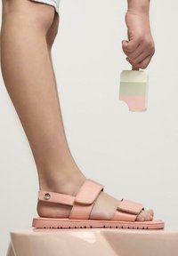 Sandalias rosas con correas ajustables y suela texturizada, acompañadas de una mano que sostiene un palito de helado de madera en tres tonos.