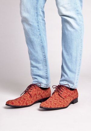 Rode schoenen met een bloemenpatroon, gemaakt van gestructureerde stof, met een spitse neus en een zwarte zool, gedragen met lichtblauwe jeans.