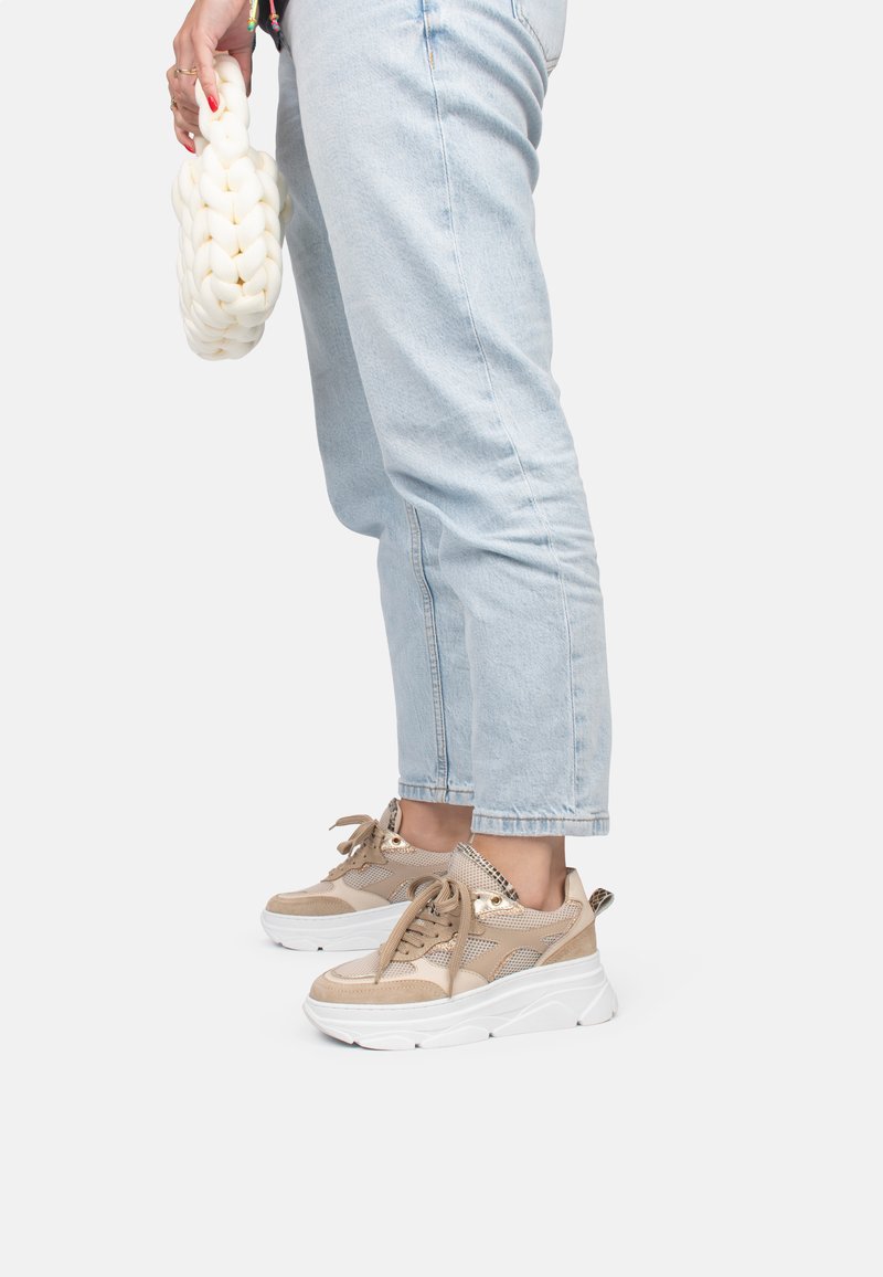 Zapatillas estilo plataforma en beige y blanco, hechas de malla y ante, con cordones redondos y detalles metálicos. Unos pantalones de mezclilla complementan el conjunto.