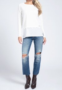 Top bianco a maniche lunghe sovrapposto a una base trasparente, abbinato a jeans blu strappati con buchi alle ginocchia e stivali con tacco in stampa rettile marrone.