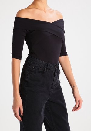 Kobieta w czarnej bluzce odkrywającej ramiona z rękawami do połowy przedramienia oraz wysokim stanem czarnych dżinsów stoi na białym tle.