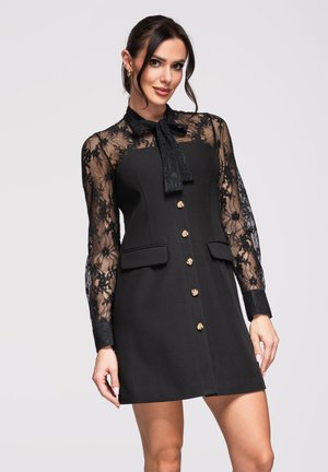 Kobieta w czarnej sukience z koronkowymi długimi rękawami, kołnierzykiem z kokardą i złotymi guzikami, stojąca na tle jasnoszarego tła.