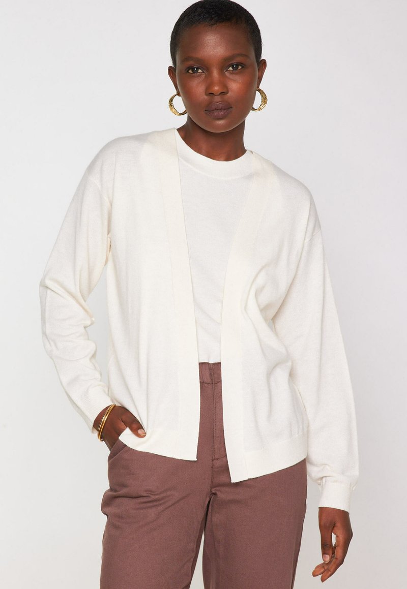 Cardigan blanc à l'avant ouvert, avec des manches longues et une texture fine, associé à un haut léger et un pantalon marron taille haute.