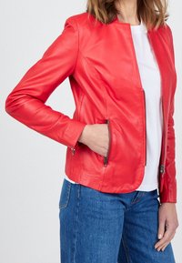 Červená kožená bunda s přiléhavým střihem, stojatým límcem a zipovými kapsami. Měkká textura a hladký povrch. V kombinaci s bílou košilí a džínami.