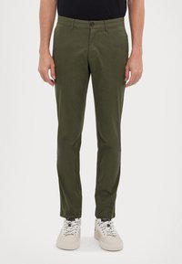 Grønne chinos laget av bomull med en jevn tekstur, med slim fit, frontlommer og knapplukking. Brukt med hvite joggesko.