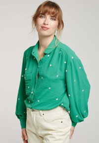 Chemise verte à boutons avec manches bouffantes, ornée de petites broderies florales blanches. Portée avec un jean de couleur claire. Tissu texturé.