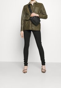 Chaqueta de cuero verde oliva con cintura ceñida, dos bolsillos frontales, combinada con leggins negros y tacones negros con tiras. Pequeño bolso negro tipo bandolera.