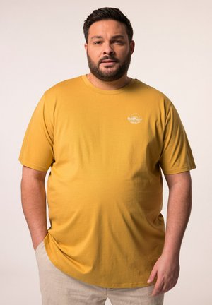 Camiseta de color mostaza en tejido suave y liso, con mangas cortas. Presenta un pequeño gráfico blanco en el lado izquierdo del pecho. Corte relajado.