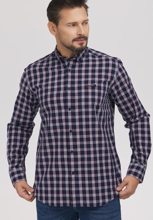 Mężczyzna w granatowo-biało-czerwonej koszuli w kratę z guzikami, poprawiający dół koszuli, patrzący na bok na jednolitym tle.