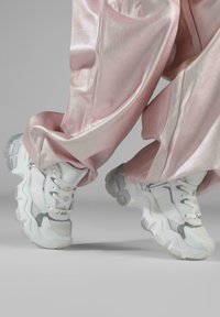Hvite klumpete sneakers med grå detaljer, mesh- og skinnmaterialer, buede såler og tykke snører, kombinert med glinsende rosa buksemateriale.