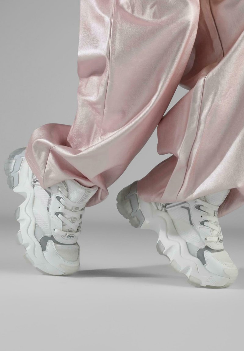 Hvite klumpete sneakers med grå detaljer, mesh- og skinnmaterialer, buede såler og tykke snører, kombinert med glinsende rosa buksemateriale.
